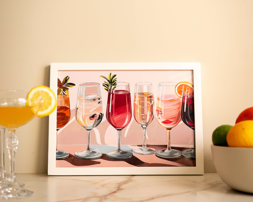 Een poster met cocktailglazen erop met drankjes erin. De poster heeft een witte lijst eromheen. Aan de linkerkant zie je een glas met een oranje drankje erin, met een sinaasappel eraan. Aan de rechterkant een fruitmand.