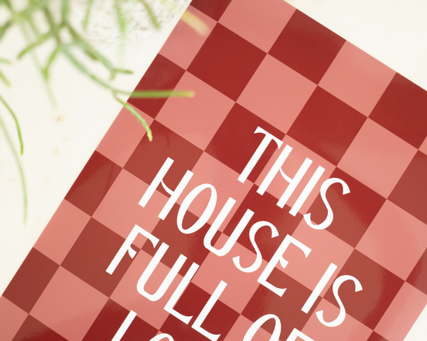 Een poster met rode en roze blokjes op de achtergrond. Op de voorgrond een witte tekst 'This house is full of love'