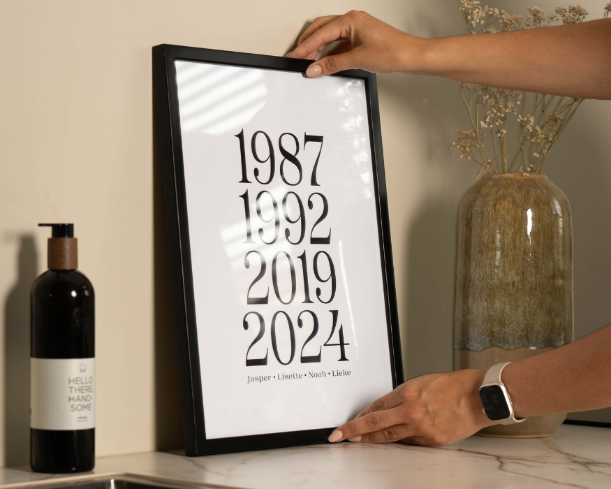 Twee handen plaatsen een ingelijste, gepersonaliseerde familieposter op een marmerkleurig oppervlak tegen een zandkleurige muur. In de strakke zwarte lijst zit een witte poster met vier grote, verticaal gestapelde jaartallen in een elegant, zwart lettertype: '1987', '1992', '2019' en '2024'. Helemaal onderaan staan de voornamen van het gezin: 'Jasper • Lisette • Noah • Lieke'. Links van de lijst staat een donkere zeepdispenser met een wit etiket waarop 'HELLO THERE HANDSOME' te lezen is. Aan de rechterkant staat een robuuste, aards getinte geglazuurde vaas met subtiele droogbloemen erin. De persoon die de lijst vasthoudt, draagt een smartwatch met een lichtgekleurd bandje om de pols.