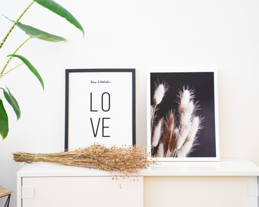 Een lichte, moderne interieuropstelling met twee ingelijste posters staand op een strakke witte kast tegen een helderwitte muur. De linker poster, in een zwarte lijst, is een gepersonaliseerd typografisch ontwerp. Bovenaan staan in een klein, sierlijk lettertype de namen 'Jurre & Natasha', met daaronder groot het woord 'LOVE' in strakke zwarte letters, speels verdeeld over twee regels ('LO' boven 'VE'). De rechter poster, in een witte lijst, toont een sfeervolle fotoprint van zachte, lichte pluimgrassen tegen een donkere achtergrond. Voor de lijsten ligt horizontaal een bosje gedroogd vlas op de kast. Aan de linkerkant buigen de bladeren van een groene kamerplant met een opvallende gestreepte stengel in beeld.