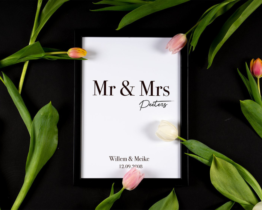 Een sfeervolle 'flatlay' foto op een effen zwarte achtergrond. In het midden ligt een gepersonaliseerde huwelijksposter in een strakke zwarte lijst. Op de witte poster staat in grote, klassieke donkere letters 'Mr & Mrs', met daaronder in een sierlijk, handgeschreven lettertype de achternaam 'Peeters'. Onderaan de poster staan de voornamen 'Willem & Meike' met daaronder de trouwdatum '12.09.2008'. De lijst wordt sierlijk omringd door verse tulpen in zachte roze, witte en geeloranje tinten, waarvan de heldergroene stengels en grote bladeren prachtig afsteken tegen de donkere ondergrond. Enkele bloemknoppen overlappen de rand van de fotolijst.