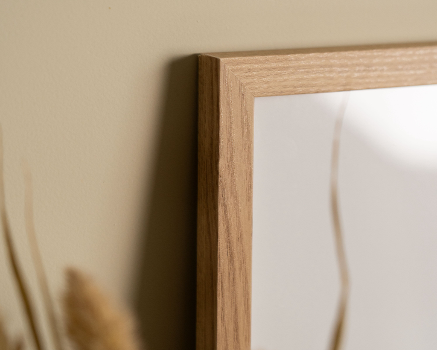 Een close-up detailopname van de linkerbovenhoek van een strakke, licht houten fotolijst (met zichtbare houtnerf). De lijst hangt of staat tegen een effen zandkleurige muur en werpt een zachte schaduw naar links. In de lijst is door de weerspiegeling van het glas een klein deel van een poster te zien met een witte achtergrond en een subtiele, organische bruine lijn. Linksonder in de wazige voorgrond steken pluimen van gedroogd gras of pampasgras in beeld.