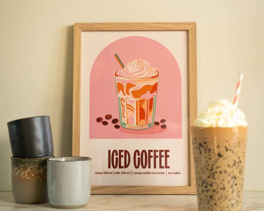 Een sfeervolle keukenopstelling met een ingelijste poster, staand op een licht marmeren aanrechtblad tegen een zandkleurige muur. In de licht houten lijst zit een poster met een retro-stijl illustratie van een grote ijskoffie met slagroom en een rietje, geplaatst voor een roze kleurvlak (een boog) met getekende koffiebonen aan de onderkant. Onderaan de poster staat in opvallende bruine letters 'ICED COFFEE', met daaronder het recept: 'lungo 150 ml | milk 100 ml | 1 scoop vanilla ice cream | ice cubes'. Links van de poster staan drie kleine keramische koffiekopjes in donkergrijze, gespikkelde en lichtgrijze tinten. Rechts op de wazige voorgrond staat een echt, hoog gespikkeld glas gevuld met ijskoffie, afgetopt met een flinke toef slagroom en een roze-wit gestreept papieren rietje.