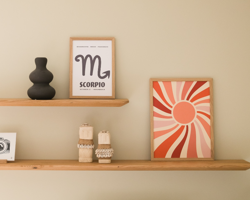 Een sfeervolle opstelling met twee licht houten wandplanken tegen een zachtbeige muur. Op de bovenste plank staat links een matzwarte, organisch gevormde vaas en rechts een poster in een houten lijst met het sterrenbeeld Schorpioen (inclusief het donkergrijze symbool, de tekst 'SCORPIO' en de bijbehorende data). Op de onderste plank staat uiterst links een klein kaartje met een illustratie van een vintage fototoestel. In het midden staan twee decoratieve, abstracte beeldjes die versierd zijn met touw en witte schelpjes. Aan de rechterkant staat een grotere poster, eveneens in een licht houten lijst, met een vrolijk, retro zonnestralen-patroon van golvende lijnen in warme tinten zoals roze, oranje, rood en crème.