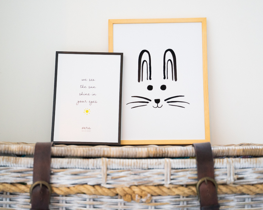 Twee ingelijste posters, staand op een wit met naturel geweven rieten kist met stoere bruine leren riemen en gespen, tegen een strakke witte muur. De linker, iets kleinere poster zit in een zwarte lijst en bevat de Engelse tekst 'we see the sun shine in your eyes' in een speels, dun lettertype. Daaronder staat een klein, vrolijk geel zonnetje getekend, gevolgd door de gepersonaliseerde naam 'vera' en de datum '02.05.2020'. De rechter, grotere poster zit in een licht houten lijst en toont een schattige, minimalistische zwarte lijntekening van een konijnengezichtje met twee lange, opstaande oren, ronde kraaloogjes en lange snorharen.
