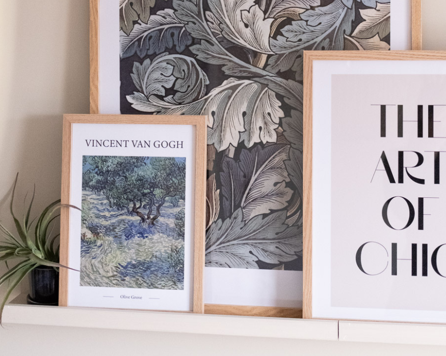 Een sfeervolle, gelaagde opstelling van drie posters in licht houten lijsten, staand op een witte wandplank tegen een zachtbeige muur. Vooraan staat een kleinere poster met een reproductie van een schilderij van Vincent van Gogh, gekenmerkt door zijn typische wervelende penseelstreken in groen- en blauwtinten. Boven het kunstwerk staat in strakke letters 'VINCENT VAN GOGH' en eronder klein 'Olive Grove'. Daarachter staat een grotere poster met een donker, weelderig botanisch bladerpatroon in de stijl van William Morris. Aan de rechterkant overlapt een lichte poster waarvan een deel van de grote, elegante zwarte typografie zichtbaar is: 'THE ART OF CHIC'. Helemaal links op de plank staat ter decoratie een klein donkerblauw potje met een groen luchtplantje.