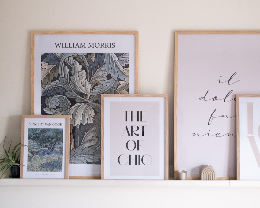 Een sfeervolle collage van ingelijste posters in licht houten lijsten, staand op een witte wandplank tegen een zachtbeige muur. Links vooraan staat een kleine poster met een reproductie van Vincent van Gogh ('Olive Grove'). Daarachter staat een grote poster met een weelderig, donker botanisch bladerpatroon in groen- en blauwtinten met de tekst 'WILLIAM MORRIS' bovenaan. In het midden overlapt een poster met de strakke zwarte typografie 'THE ART OF CHIC'. Aan de rechterkant staat een grote zachtroze poster met de sierlijke, cursieve tekst 'il dolce far niente', deels overlapt door nog een andere lichte poster. Ter decoratie staat uiterst links een klein luchtplantje in een donkerblauw potje, en rechts voor de lijsten staan een klein zilverkleurig theelichthoudertje en een decoratieve beige, boogvormige ribbelkaars.