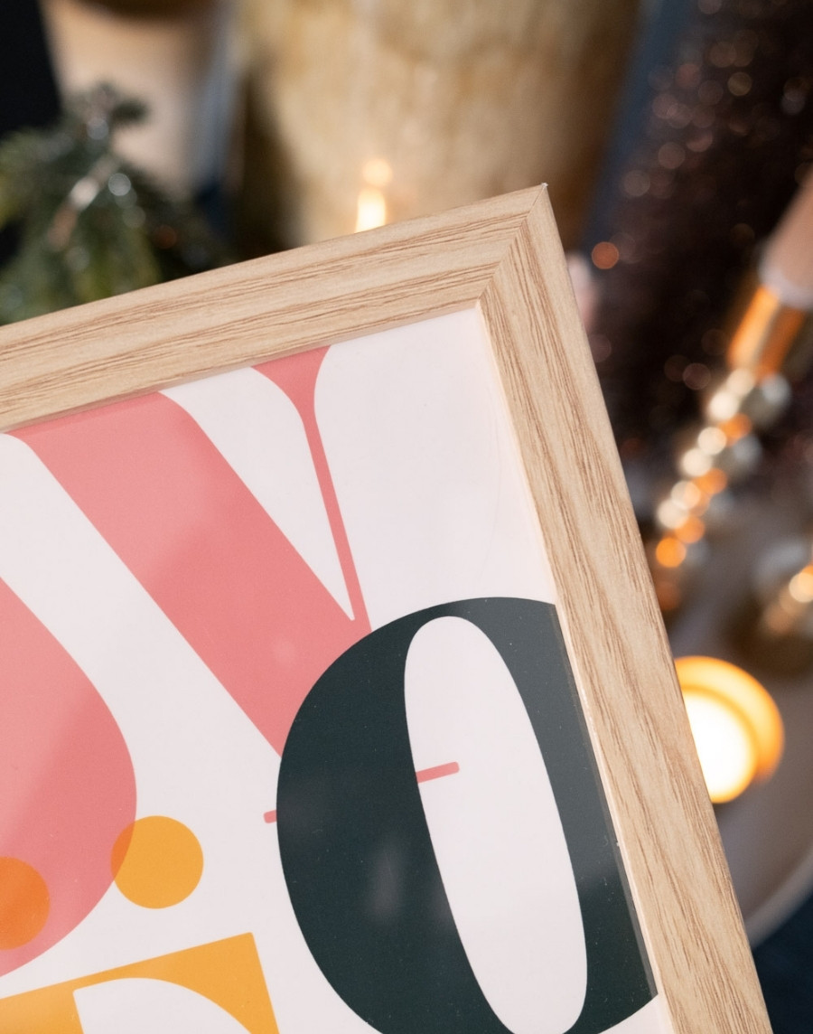 Close-up van de rechterbovenhoek van een houten fotolijst met een kleurrijke kerstposter met grafische letters, tegen een sfeervol vervagende achtergrond met warme kerstverlichting.