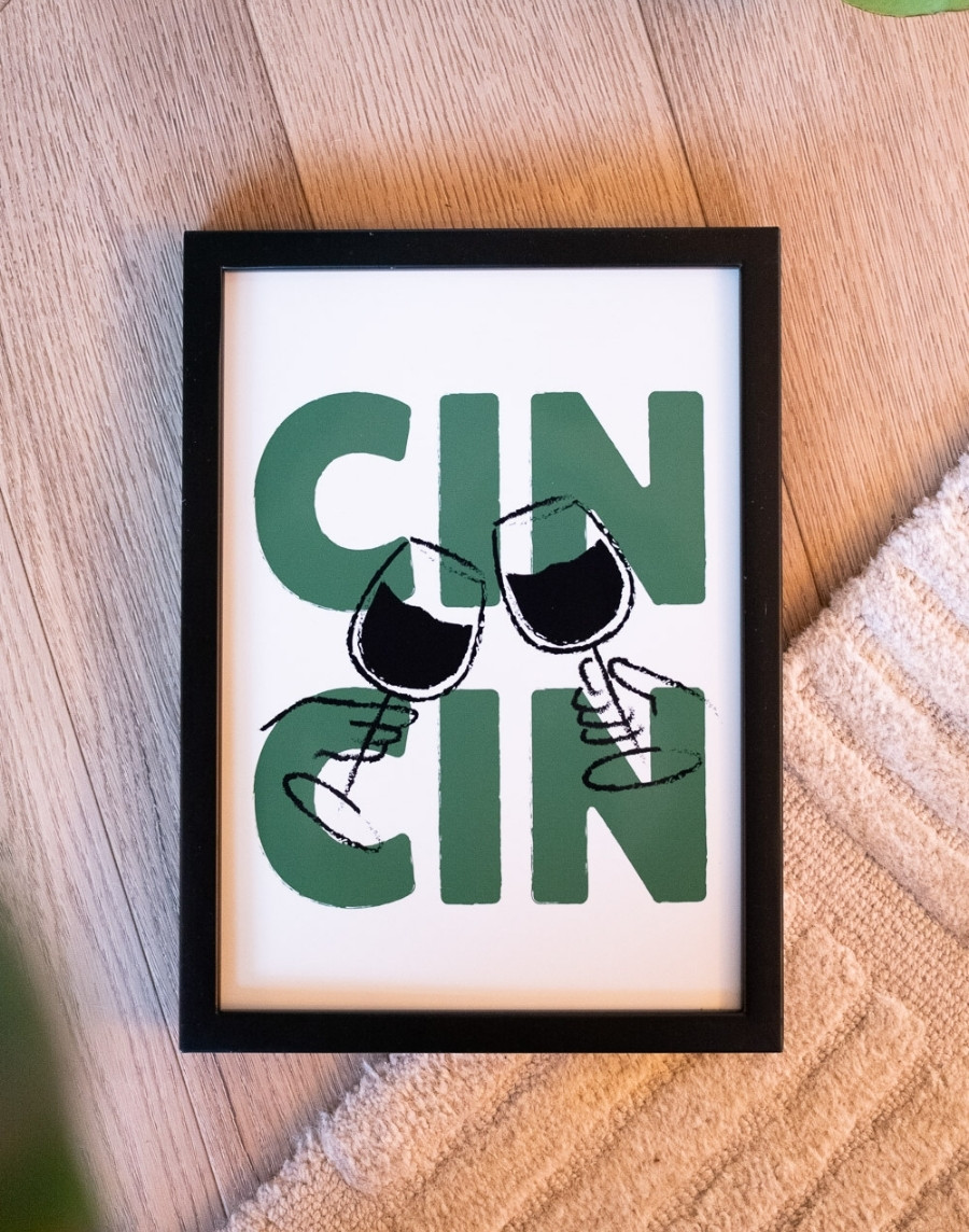 Bovenaanzicht van een ingelijste 'Cin Cin' poster met groene letters en proostende wijnglazen in een zwarte lijst, liggend op een lichte houten vloer naast een harig tapijt.