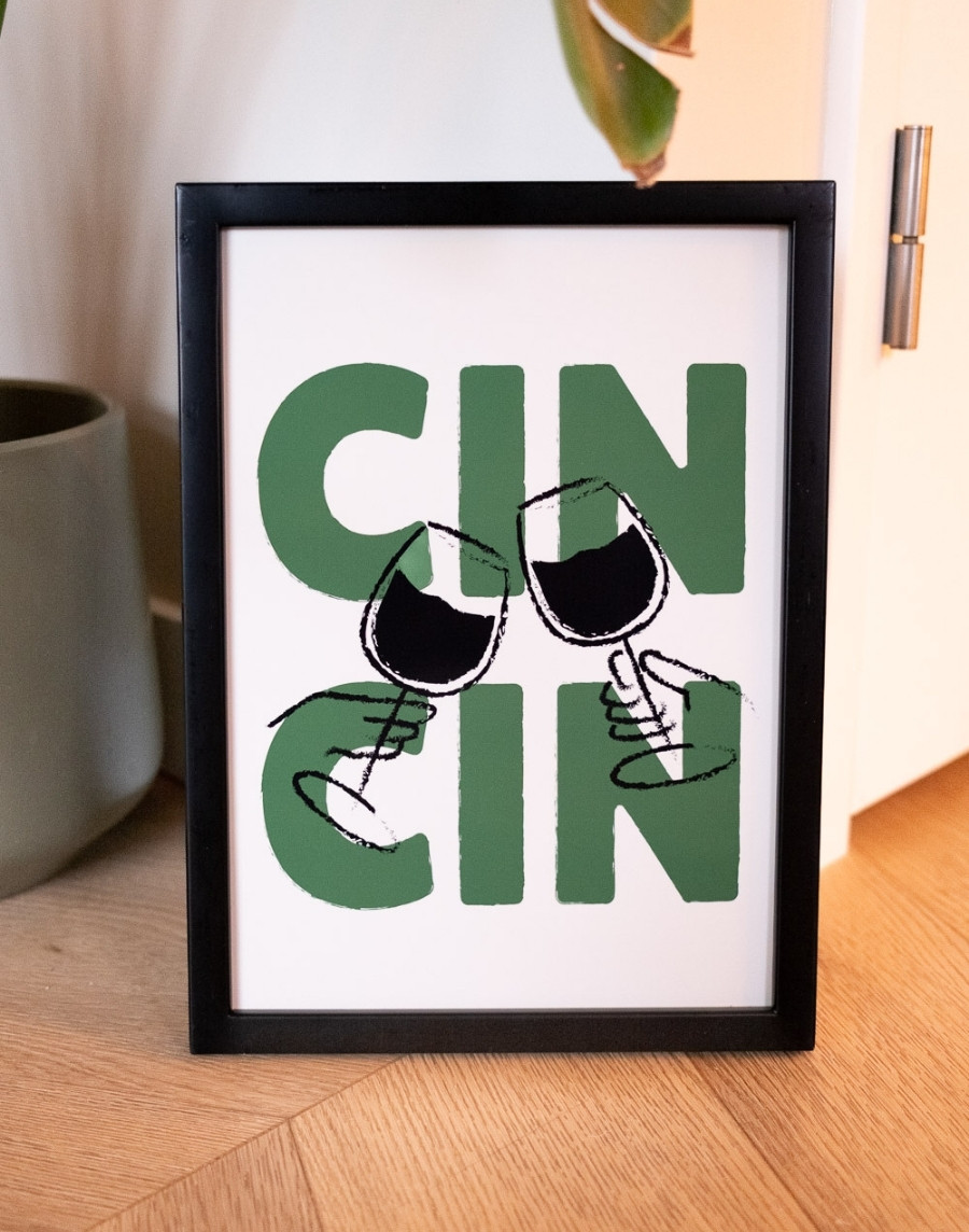 Ingelijste poster met de tekst 'Cin Cin' in groene letters en een illustratie van twee proostende wijnglazen in een zwarte fotolijst, staand op een houten visgraatvloer.