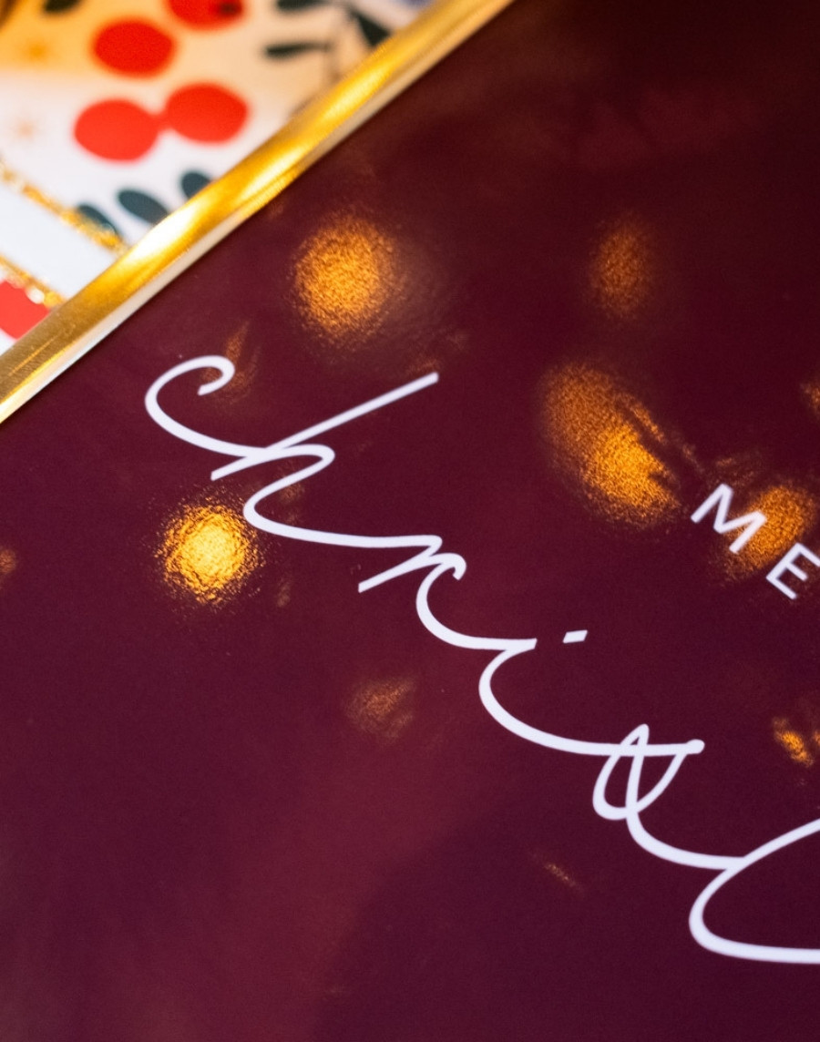 Close-up van een ingelijste kerstposter met witte sierletters op een dieprode achtergrond in een gouden fotolijst, met sfeervolle lichtreflecties.