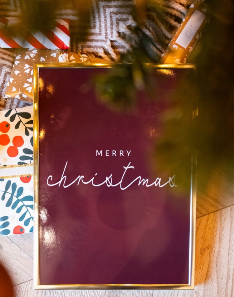 Bovenaanzicht van een ingelijste kerstposter met de tekst 'MERRY christmas' op een donkerrode achtergrond in een gouden fotolijst, liggend tussen ingepakte kerstcadeaus onder de kerstboom.