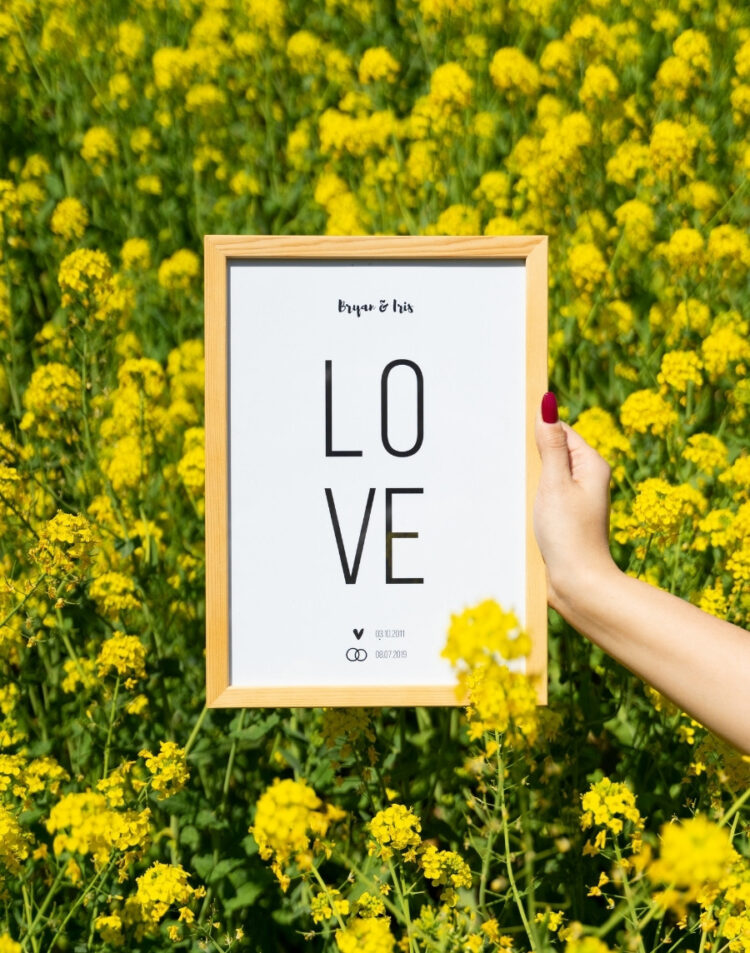 Hand die een ingelijste poster vasthoudt met de tekst 'LOVE', de namen Bryan & Iris en twee data met symbolen, in een houten lijst tegen een achtergrond van een veld vol gele bloemen.