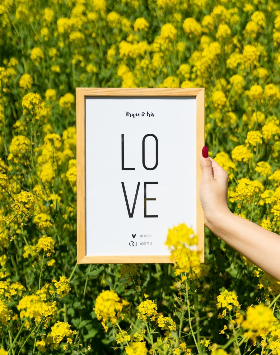 Hand die een ingelijste poster vasthoudt met de tekst 'LOVE', de namen Bryan & Iris en twee data met symbolen, in een houten lijst tegen een achtergrond van een veld vol gele bloemen.