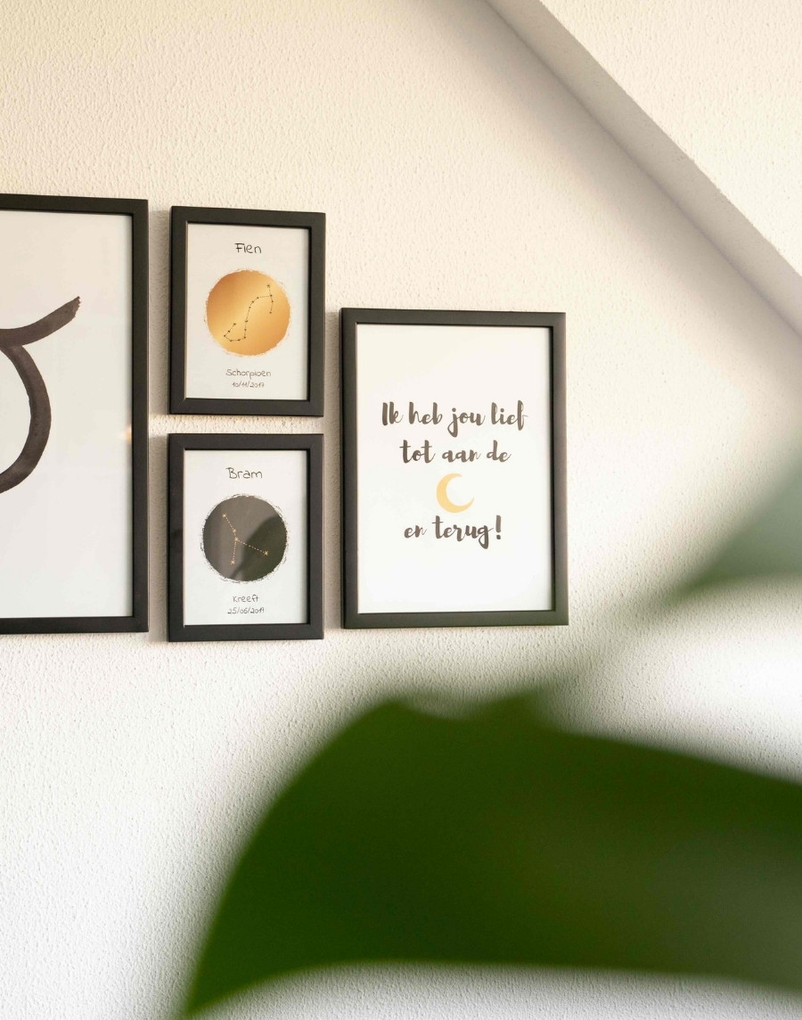 Fotowand met zwarte fotolijsten op een witte muur, bestaand uit twee gepersonaliseerde sterrenbeeldposters voor Fien en Bram en een poster met de tekst 'Ik heb jou lief tot aan de maan en terug!'.