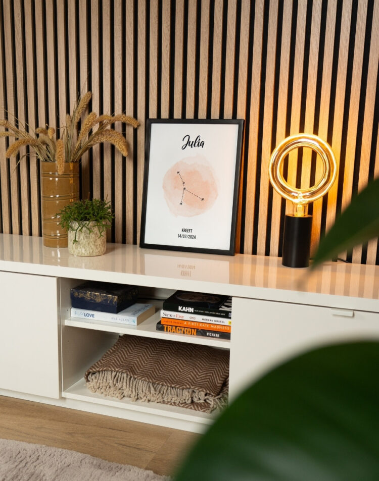 Ingelijste poster met het sterrenbeeld Kreeft en de naam Julia op een wit tv-meubel, gestyled tegen een akupanel lattenwand met een design lamp, planten en woondecoratie.