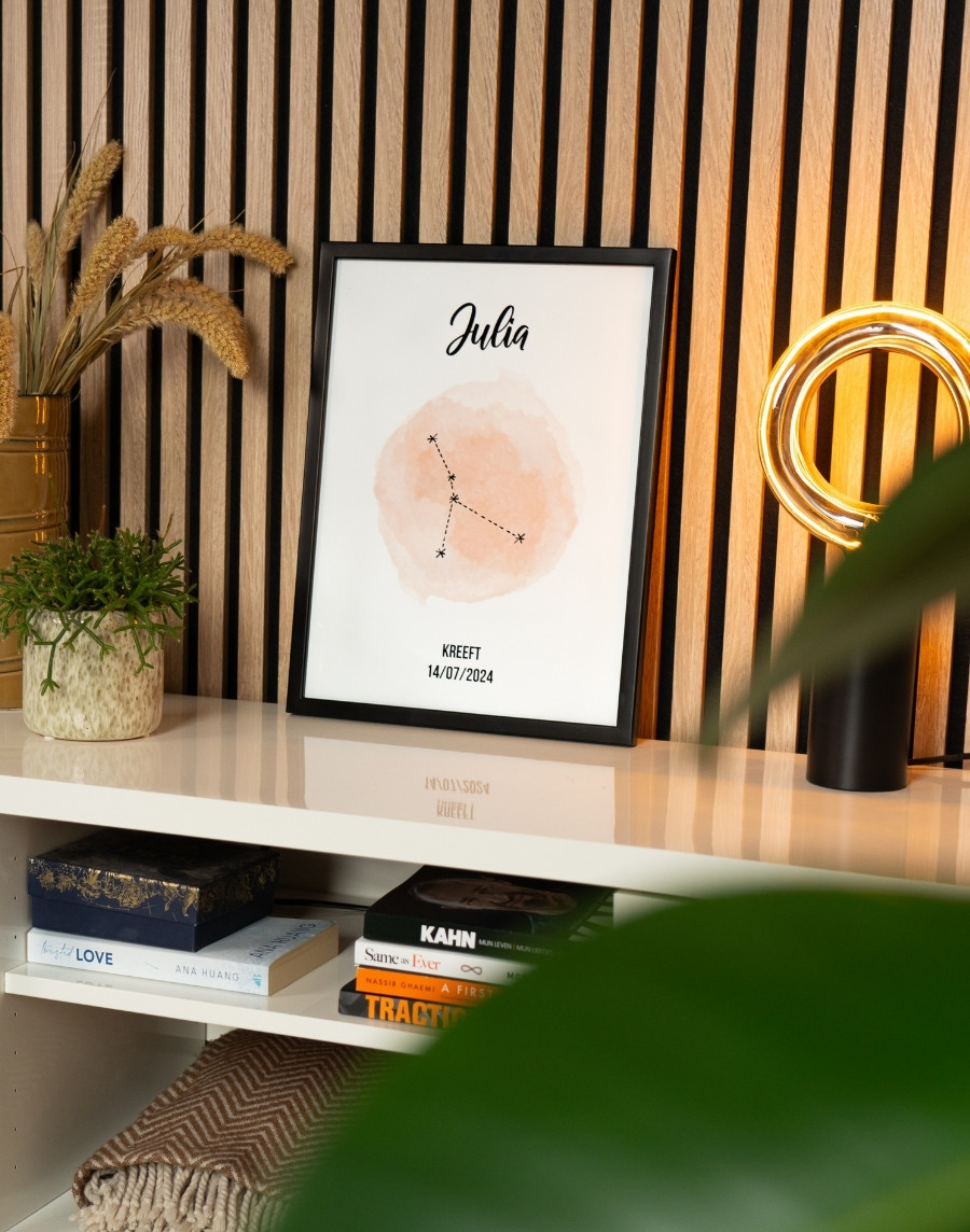 Close-up van een ingelijste kinderposter met het sterrenbeeld Kreeft en de naam Julia, in een zwarte fotolijst op een wit dressoir tegen een houten lattenwand.