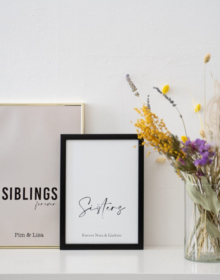 Twee gepersonaliseerde posters met de teksten 'Siblings forever Pim & Lisa' in een gouden lijst en 'Sisters Forever Nora & Liselotte' in een zwarte fotolijst, gestyled naast een vaas met kleurrijke droogbloemen.