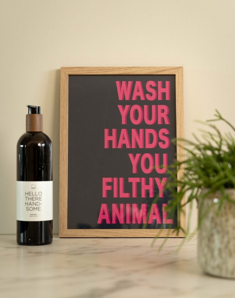 Ingelijste badkamerposter met de roze tekst 'Wash Your Hands You Filthy Animal' in een houten fotolijst, gestyled naast een fles zeep en een groen plantje op een marmeren planchet.