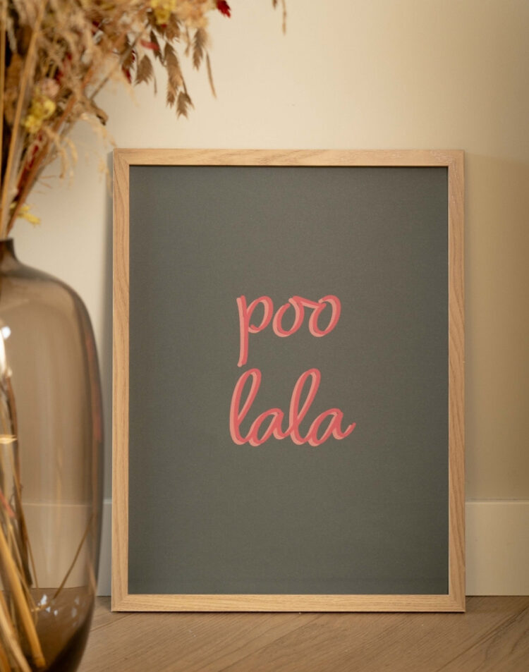 Ingelijste typografie toiletposter met de roze tekst 'Poo Lala' op een donkergrijze achtergrond in een houten lijst, gestyled naast een vaas met droogbloemen.