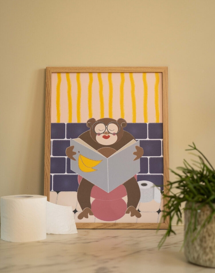 Ingelijste toiletposter met een grappige illustratie van een aap met bril die de krant leest op het toilet in een houten lijst, gestyled naast een rol toiletpapier en een plantje.