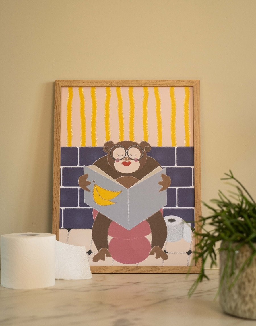 Ingelijste toiletposter met een grappige illustratie van een aap met bril die de krant leest op het toilet in een houten lijst, gestyled naast een rol toiletpapier en een plantje.