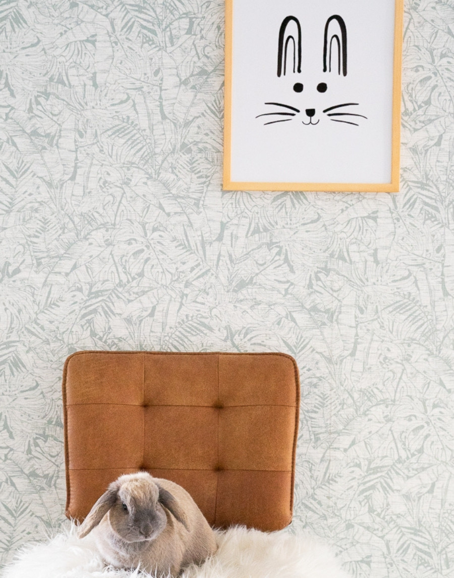 Sfeerbeeld van een ingelijste kinderposter met een konijnensnuit in een houten lijst aan een muur met bladerenbehang, boven een cognackleurige stoel met een wit bontkleed en een dwergkonijn.