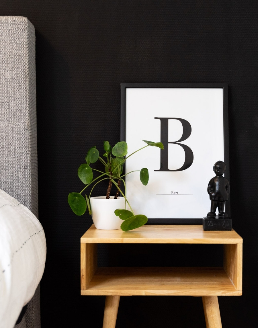 Ingelijste typografie poster met de letter B en de naam Bart in een zwarte fotolijst, gestyled op een houten nachtkastje naast een pannenkoekplant en een zwart beeldje.
