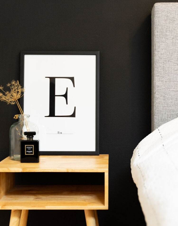 Ingelijste typografie poster met de letter E en de naam Eva in een schwarze fotolijst, gestyled op een houten nachtkastje naast een bed met een flesje parfum en droogbloemen.