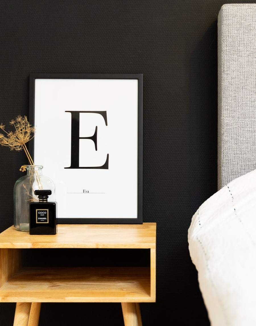 Ingelijste typografie poster met de letter E en de naam Eva in een schwarze fotolijst, gestyled op een houten nachtkastje naast een bed met een flesje parfum en droogbloemen.