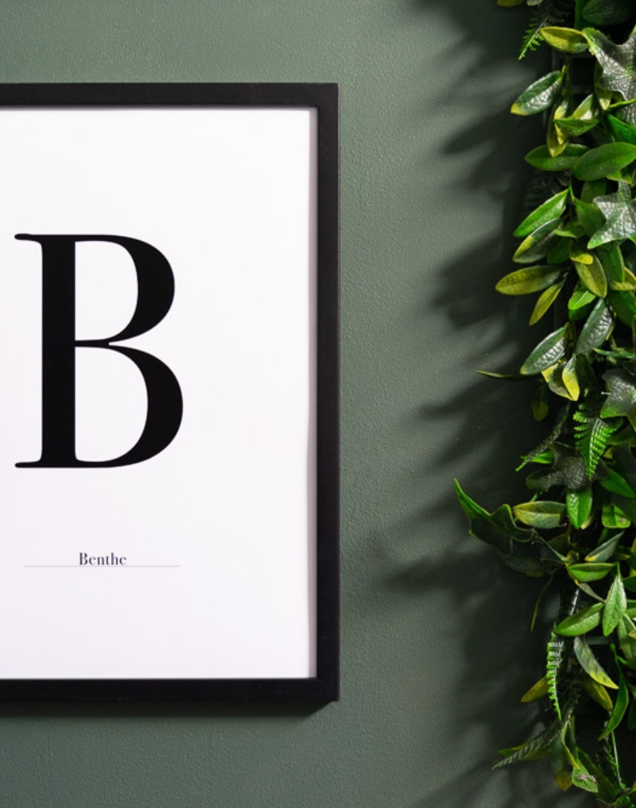 Close-up van een ingelijste typografie poster met de letter B en de naam Benthe in een zwarte lijst op een groene muur naast een kunstplant.