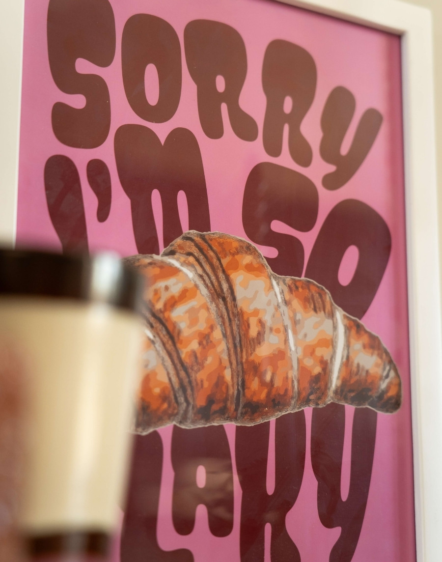 Gedetailleerde close-up van een ingelijste roze poster met een croissant en de tekst 'Sorry I'm So Flaky' in een witte fotolijst met een onscherpe beker op de voorgrond.