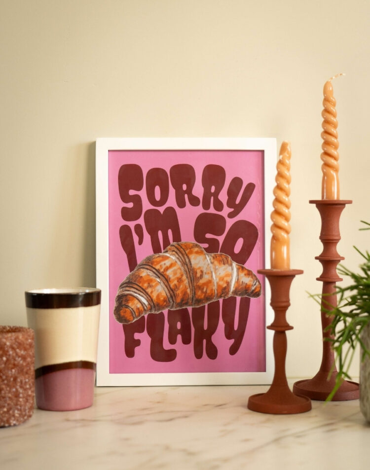Close-up van een ingelijste roze poster met een croissant en de tekst 'Sorry I'm So Flaky' in een witte fotolijst, gestyled naast terra kandelaars en een keramieken beker.