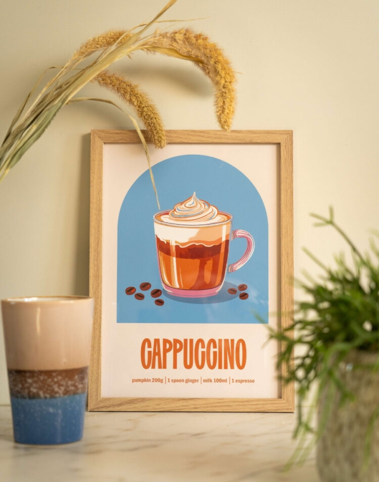 Ingelijste keukenposter met een illustratie van een cappuccino en recept op een blauwe achtergrond in een houten lijst, gestyled met droogbloemen, een koffiebeker en een kamerplant.