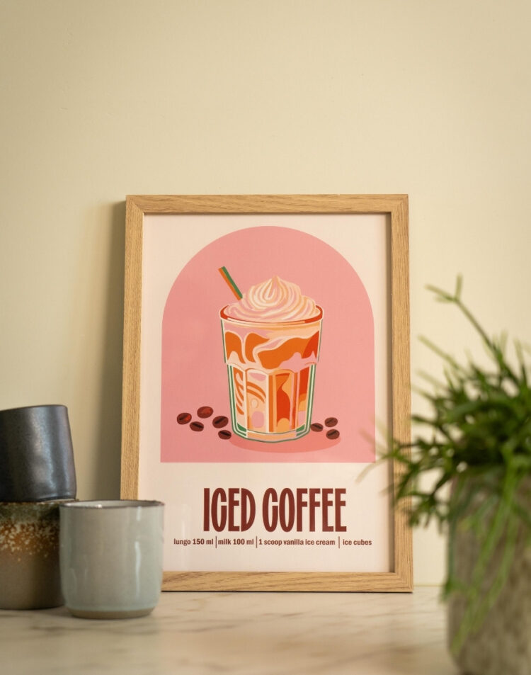 Close-up van een ingelijste poster met een illustratie van iced coffee en recept in een houten lijst, gestyled op een aanrecht naast koffiemokken en een kamerplant.