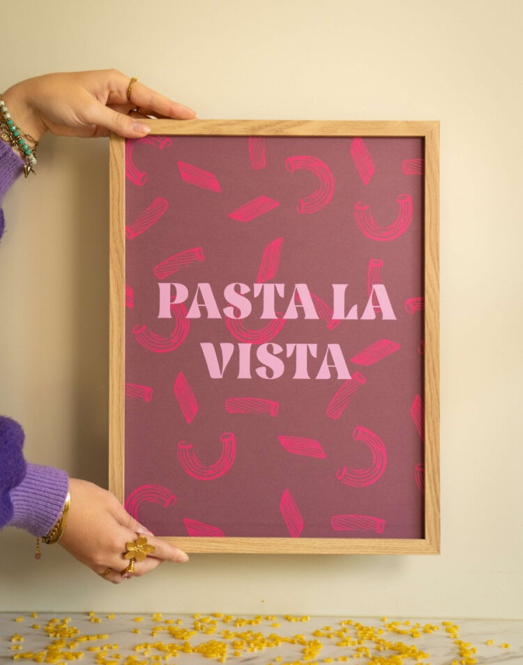 Frontale opname van een roze ingelijste poster met de tekst 'Pasta la Vista' in een houten lijst, vastgehouden door iemand met een paarse trui boven een met pasta besprenkelde ondergrond.