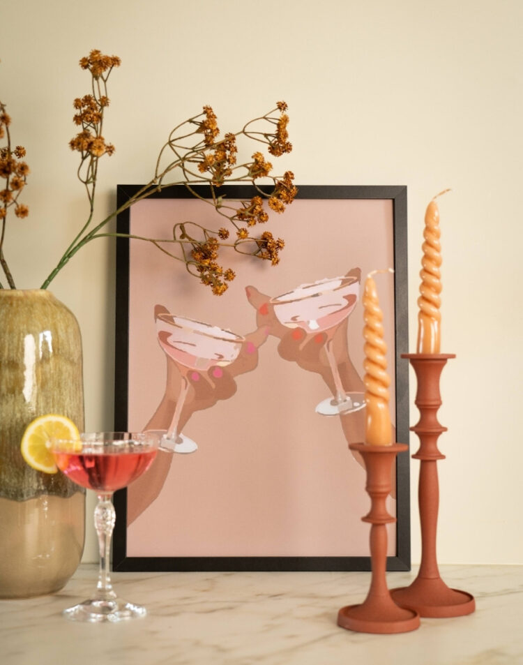 Ingelijste poster met een illustratie van twee handen die proosten met cocktailglazen in een schwarze fotolijst, gestyled op een tafel met kandelaars, droogbloemen en een roze cocktail.