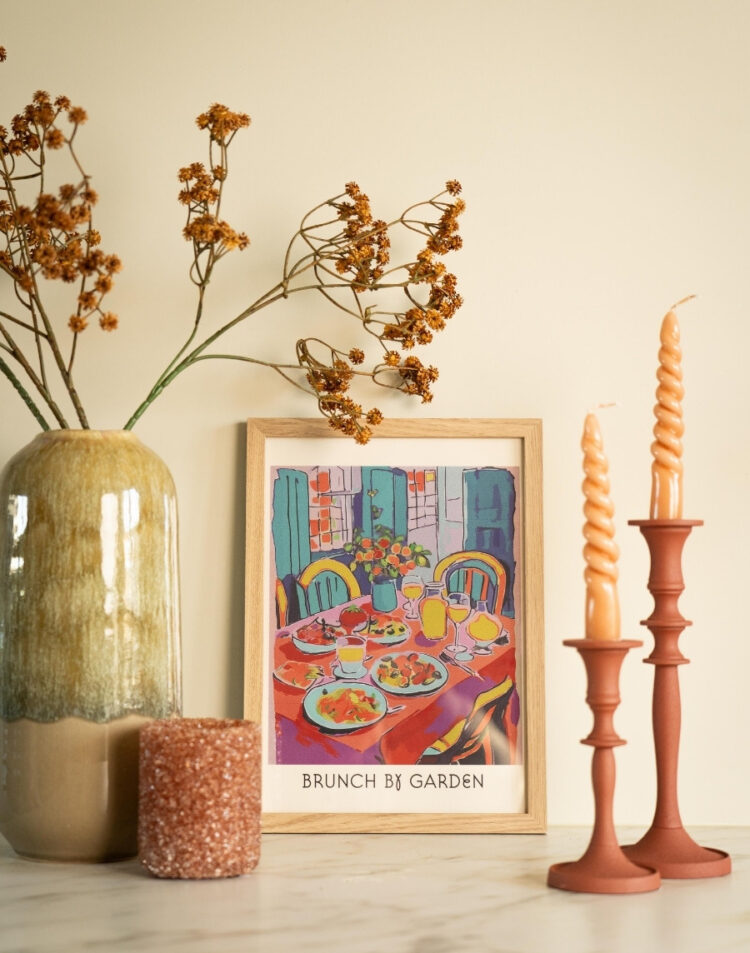 Ingelijste kunstposter met een kleurrijke illustratie van een gedekte tafel en de tekst 'Brunch by Garden' in een houten lijst, gestyled met kaarsen en een vaas droogbloemen.