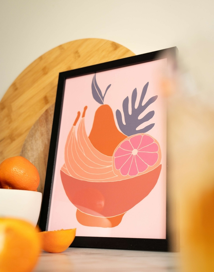 Schuinstaande ingelijste kunstposter met een illustratie van een fruitschaal in een zwarte fotolijst, geplaatst op een aanrecht met citrusvruchten en snijplanken op de achtergrond.