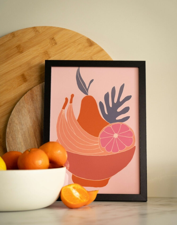 Close-up van een ingelijste kunstposter met een illustratie van een fruitschaal met bananen, peren en citrusvruchten op roze achtergrond in zwarte lijst.