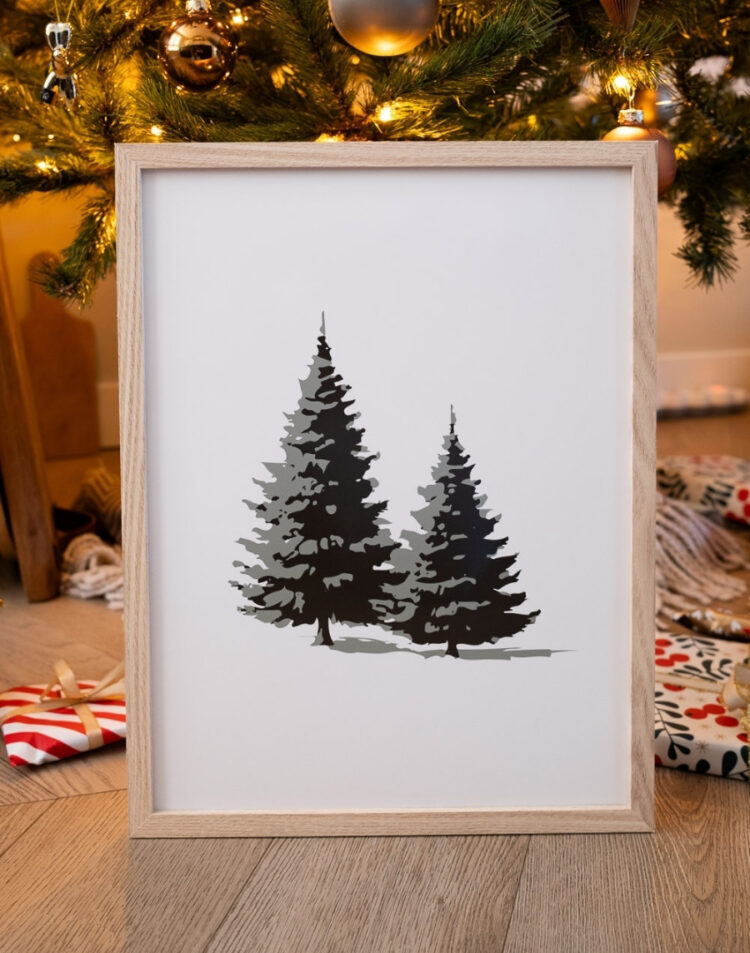 Ingelijste poster met een minimalistische illustratie van twee dennenbomen in een houten fotolijst, staand onder een versierde kerstboom op een houten vloer.