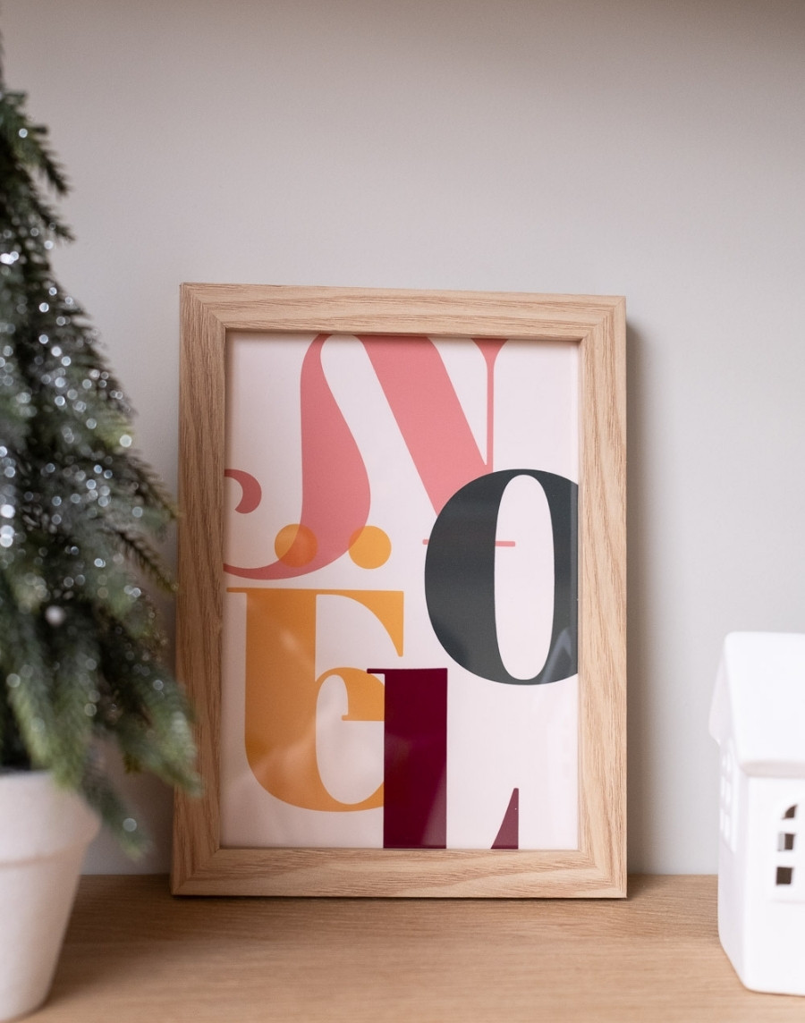 Een ingelijste, kleurrijke typografische kerstposter met de tekst 'Noël', staand op een houten plank naast een kunstmatige kerstboom in een pot en een wit keramisch huisje.