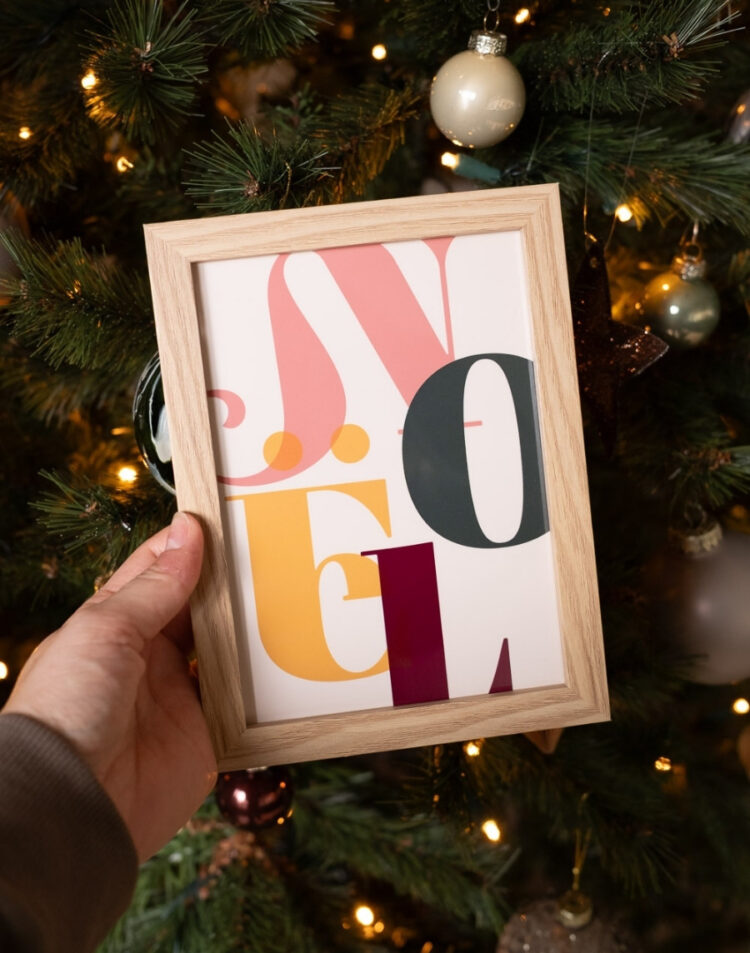 Hand die een ingelijste kerstposter met de tekst 'Noël' vasthoudt voor een verlichte kerstboom met kerstballen.