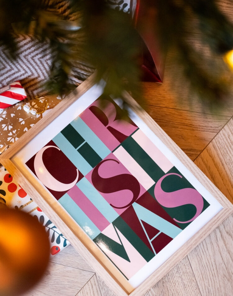 Kleurrijke kerstposter met het woord Christmas in houten lijst onder een kerstboom met cadeautjes op een houten vloer.