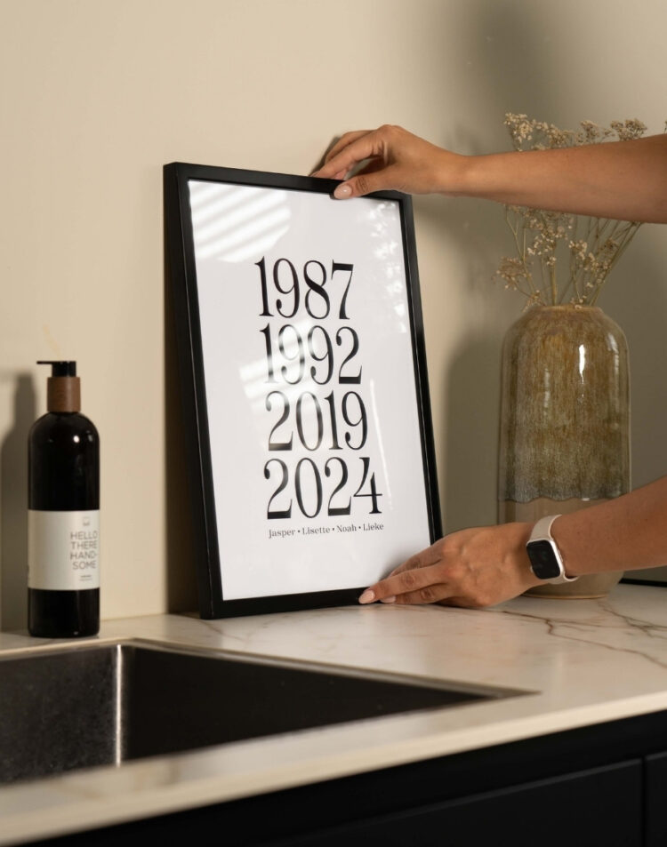 Handen die een zwarte fotolijst met gepersonaliseerde jaartallen poster neerzetten op een keukenaanrecht.