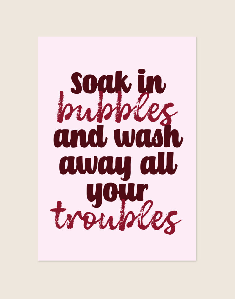 Sfeervolle lichtroze typografische poster met bordeauxrode tekst soak in bubbles and wash away all your troubles, sfeervol opgehangen aan een neutrale muur in een moderne badkamer.