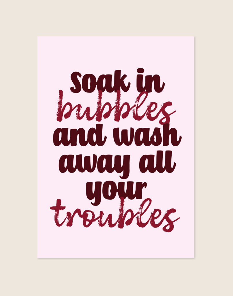 Sfeervolle lichtroze typografische poster met bordeauxrode tekst soak in bubbles and wash away all your troubles, sfeervol opgehangen aan een neutrale muur in een moderne badkamer.