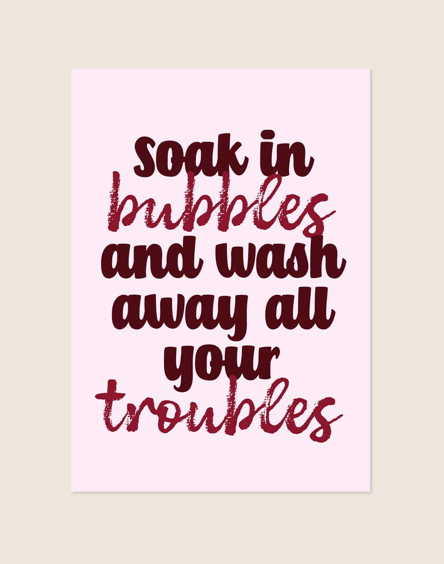 Sfeervolle lichtroze typografische poster met bordeauxrode tekst soak in bubbles and wash away all your troubles, sfeervol opgehangen aan een neutrale muur in een moderne badkamer.