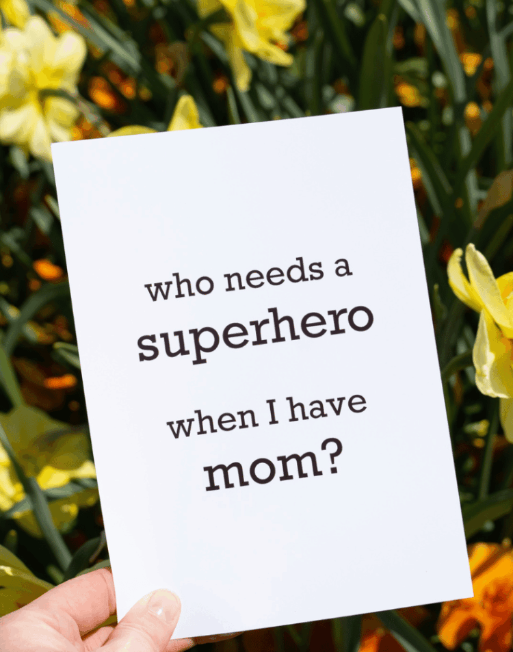 Een hand houdt een witte kaart vast tegen een achtergrond van gele narcissen en groen blad. Op de kaart staat in zwarte letters de Engelse tekst "who needs a superhero when I have mom?" gedrukt.