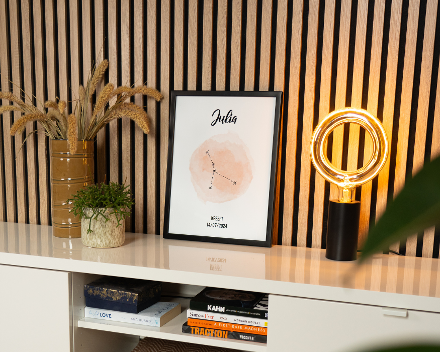 Een ingelijste gepersonaliseerde poster, staand op een glanzend wit meubel tegen een achterwand met verticale houten latten. In de strakke zwarte lijst zit een poster met de naam 'Julia' in sierlijke letters, een zachtoranje aquarel vlek met daarin de sterrenconstellatie van de Kreeft, en onderaan de tekst 'KREEFT' met de datum '14/07/2024'. Rondom de poster staan sfeervolle woonaccessoires: links een bruine vaas met gedroogde grassen en een klein groen plantje, en rechts een moderne zwarte tafellamp met een opvallende grote, ronde lichtbron. In het open vak onder de poster liggen diverse boeken opgestapeld.